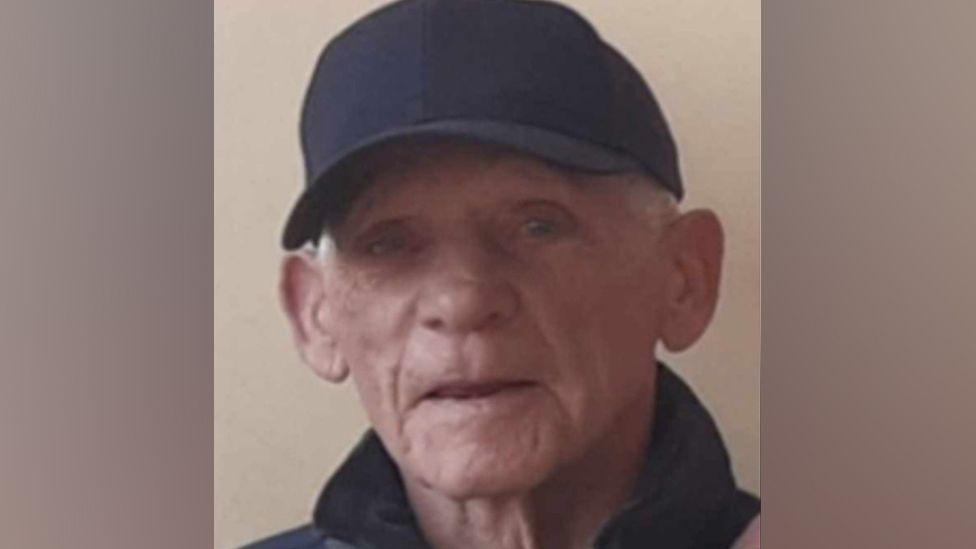 An older man with grey hair, wearing a black baseball cap and jacket.