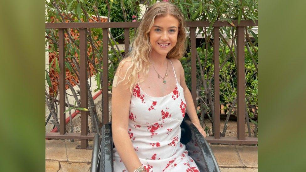 Emma Daniels, a young woman with blonde hair, is looking at the camera and smiling. She is wearing a white and red floral patterned dress. She is sitting in a wheelchair.