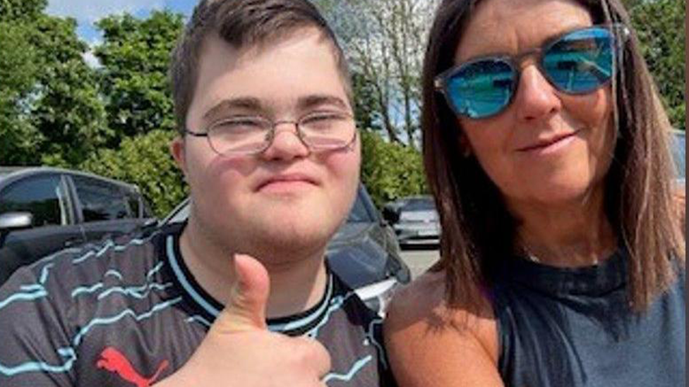 A boy in a black and blue stripy tshirt with glasses and his thumb up next to a woman in a dark blue top and reflective sunglasses