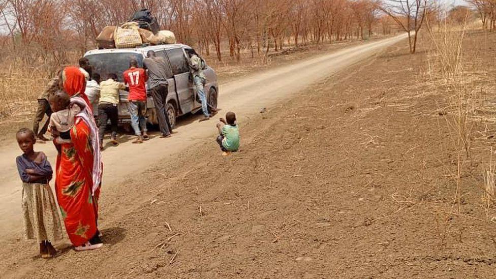 Men pushing a vehicle loaded with baggage on its roof along a dirt road in Sudan.