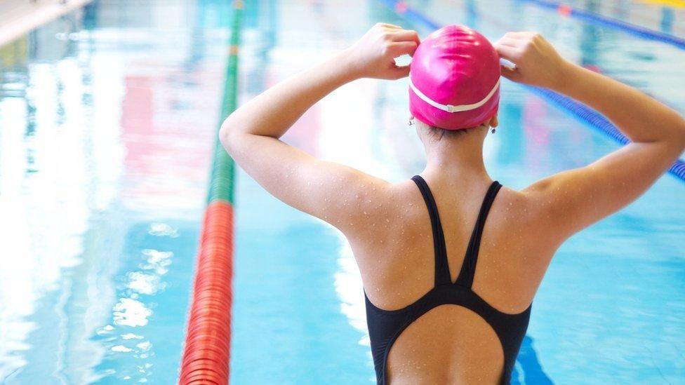 A female swimmer wearing a pink cap has her back to us with her hands at either side of her head, wearing a dark swimming costume. In front of her is a swimming pool with marked off lanes.