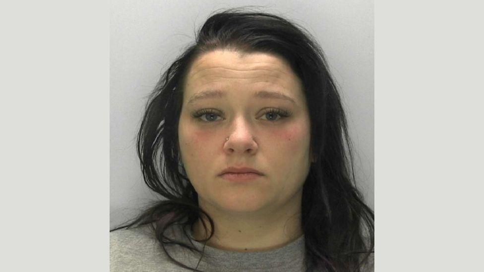 A police mugshot of Melissa Wilband wearing a grey sweatshirt and standing against a grey background. She has long dark hair in a side parting, a stud piercing in her right nostril, and eyelash extensions. She is looking at the camera with a blank expression, with red skin under her eyes.