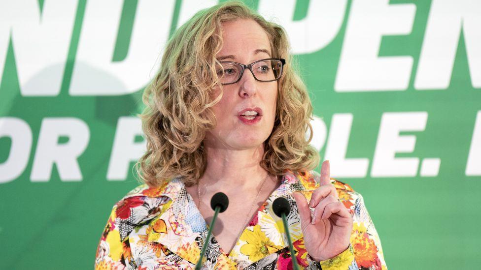 A blonde-haired woman with black glasses speaks with a raised pointed finger at a press conference with a green background 