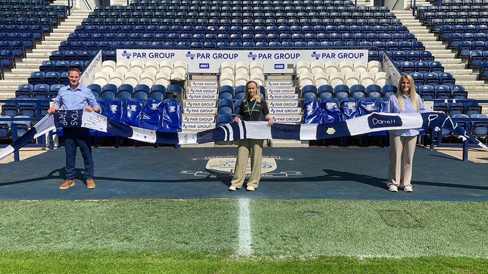 A man and two women holding the long navy blue and white striped scarf on the pitch at PNE