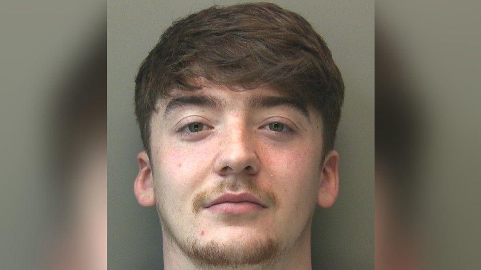 A police head-and-shoulders shot of a man with brown hair swept across his forehead and a light brown moustache and chin hair.
