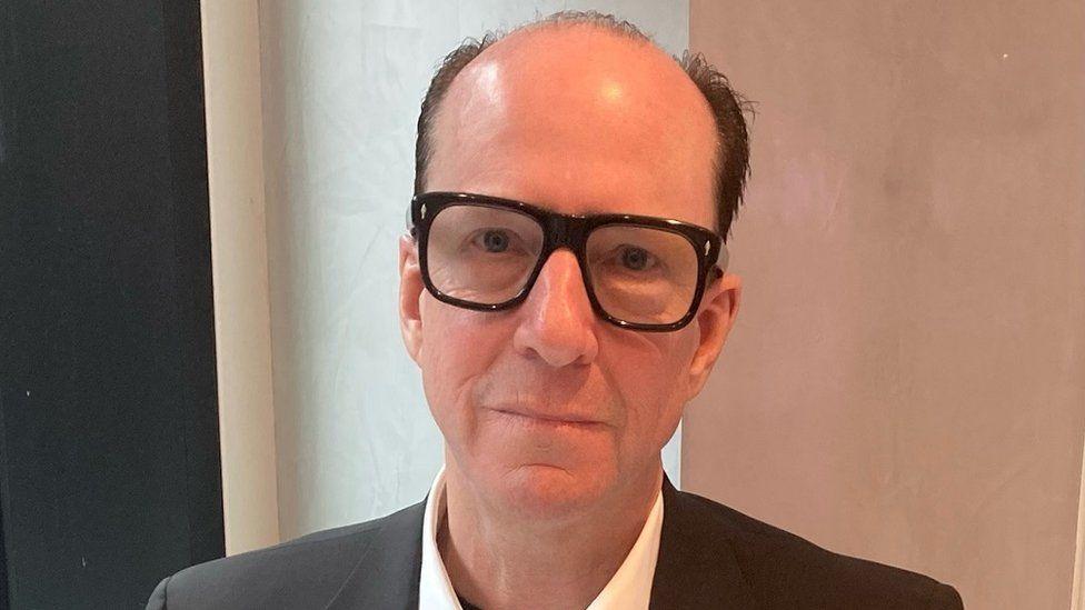 A man wearing a jacket and white shirt with dark brown rimmed glasses. He has short brown hair and is balding.