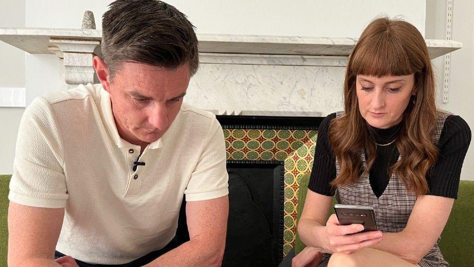 Two people sat looking down at phones. One is a man with short dark hair. He is wearing a white polo shirt. A woman with long red hair is sat next to him in a black top and checked dress.
