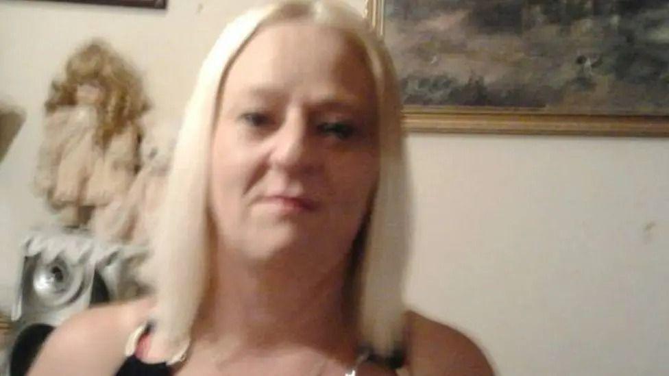 Debbie Leitch's mother Elaine Clarke. She is wearing a black top with silver loops on the shoulders. She has bleached blonde hair that is shoulder length.