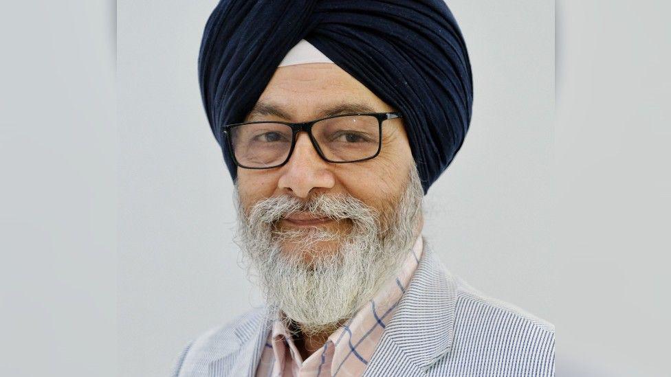 A professional headshot of a man with a grey beard, glasses and a black turban, smiling at the camera against a white background.