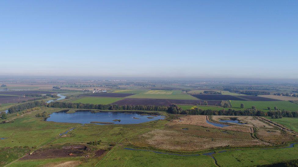 An aerial view of Kingfishers Bridge nature reserve in the summer. It shows a patchwork of fields and large and small ponds, plus a meandering stream. Beyond it are fields and above it a hazy blue sky.