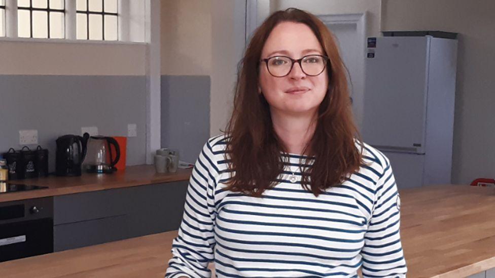 Laura White smiling in the new kitchen in front of a large wooden worktop, with a fridge and kettles on a counter top in the background