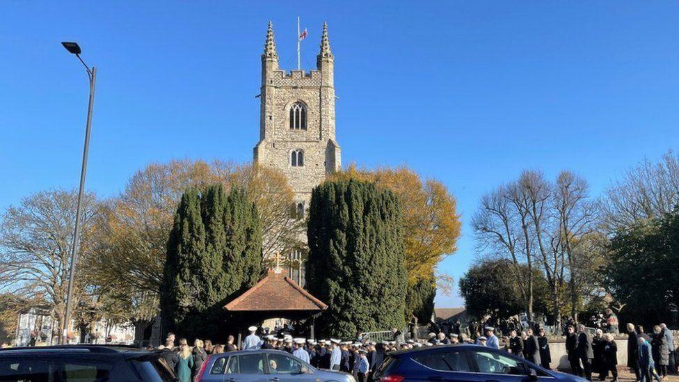 A large church with two large conifer trees outside. A St George's flag flies at half mast at the top of the church tower. Dozens of people are gathered outside the church 