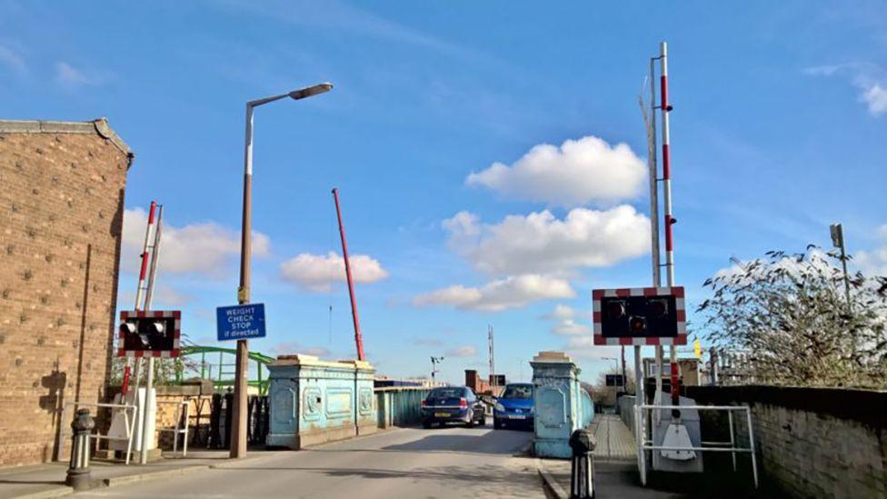 Sulcoates Bridge before its closure to traffic in 2020. It has light blue metal structure and barriers and warning lights at the end of the bridge. Two cars are driving across.