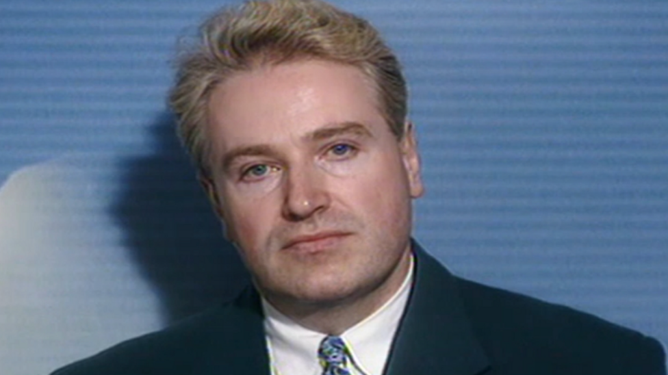 Neil Foden as a young man pictured against a blue background and wearing a dark suit and blue tie