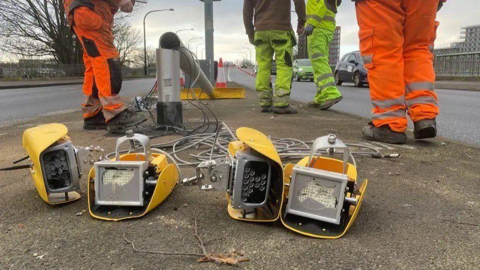 Camera in four sections with yellow casing, wires and a pole are across a road island, legs of four people in high-vis are standing by the camera.