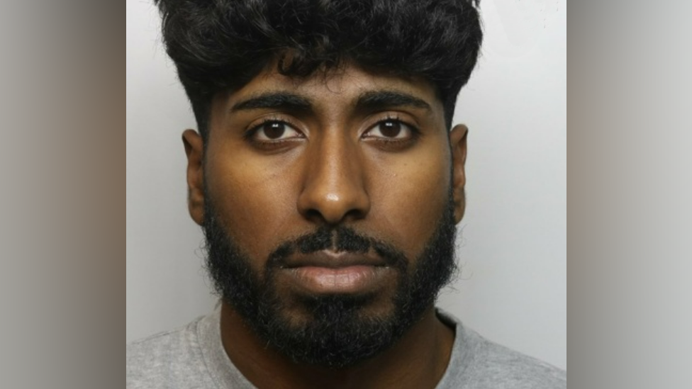 A police mugshot of a man with a beard in a light grey t-shirt