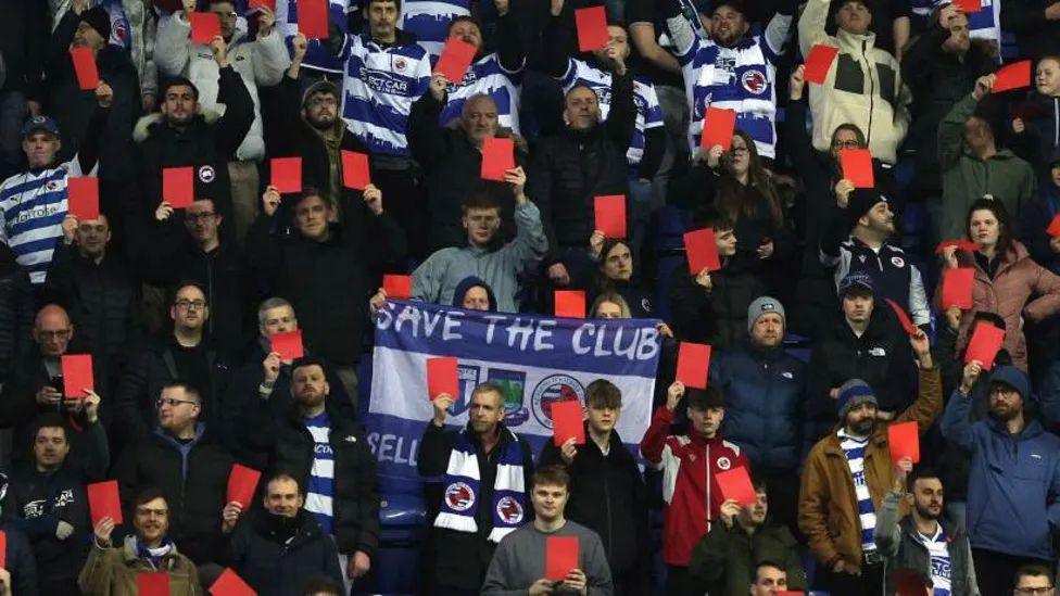 Fans in a stand, all holding large red cards, with a flag saying 'Save the club' 