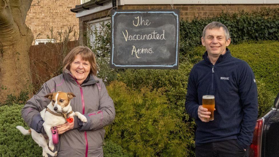 A woman, holding a dog, and a man, holding a pint of beer, stand outside in coats and smile in front of a sign that says "the Vaccinated Arms"