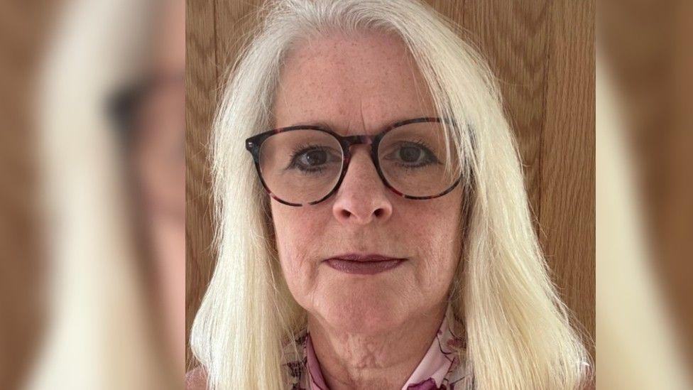 Close up of Sue's face, she has blonde hair and black glasses with brown eyes. She is stood in front of a wooden frame.
