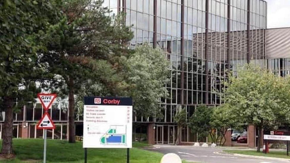 An external view of RS Components in Corby. The image shows a building with reflective glass windows standing behind trees and a sign detailing a map of the site.