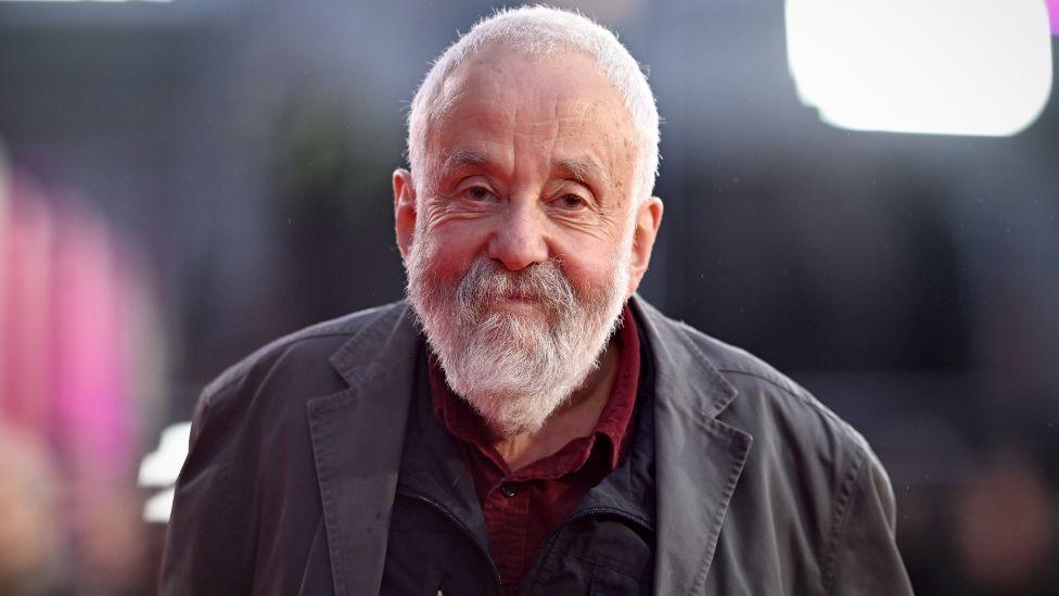 Mike Leigh looks at the camera. He has a white beard and moustache and is wearing a grey jacket and two layers underneath