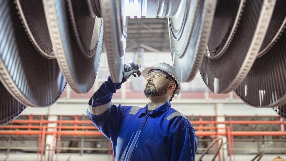 An engineer wearing blue overalls, a white hard hat and safety goggles inspects a turbine in a nuclear power station.