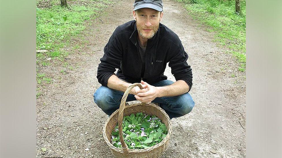 John Rensten in blue jeans, a jumper and baseball cap. He is crouched in front of a woven basket filled with green leaves.