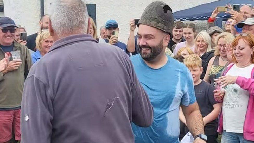 Aaron Sanderson during the coronation. He is wearing the rusty and damaged crown. He is surrounded by people watching on and smiling. His blue t-shirt is wet and he is smiling.