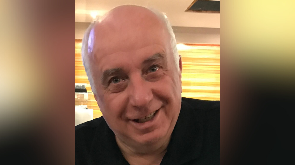 A headshot of a man smiling at the camera. He is wearing a black shirt. He has white hair around the sides of his head.
