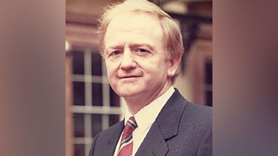 Kevin Hickey with ginger hair, white shirt, red, white and black club tie and dark grey  suit