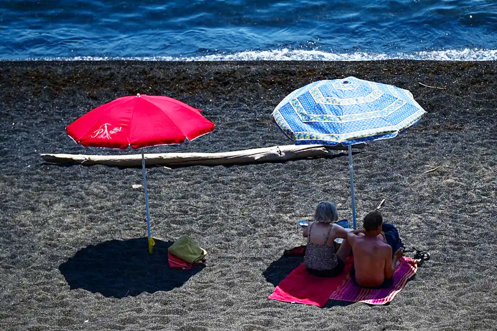 A red and blue parasol on a beach next to the sea, which is at the top of the picture. Two beachgoers are resting in the shade under the blue parasol on the right.