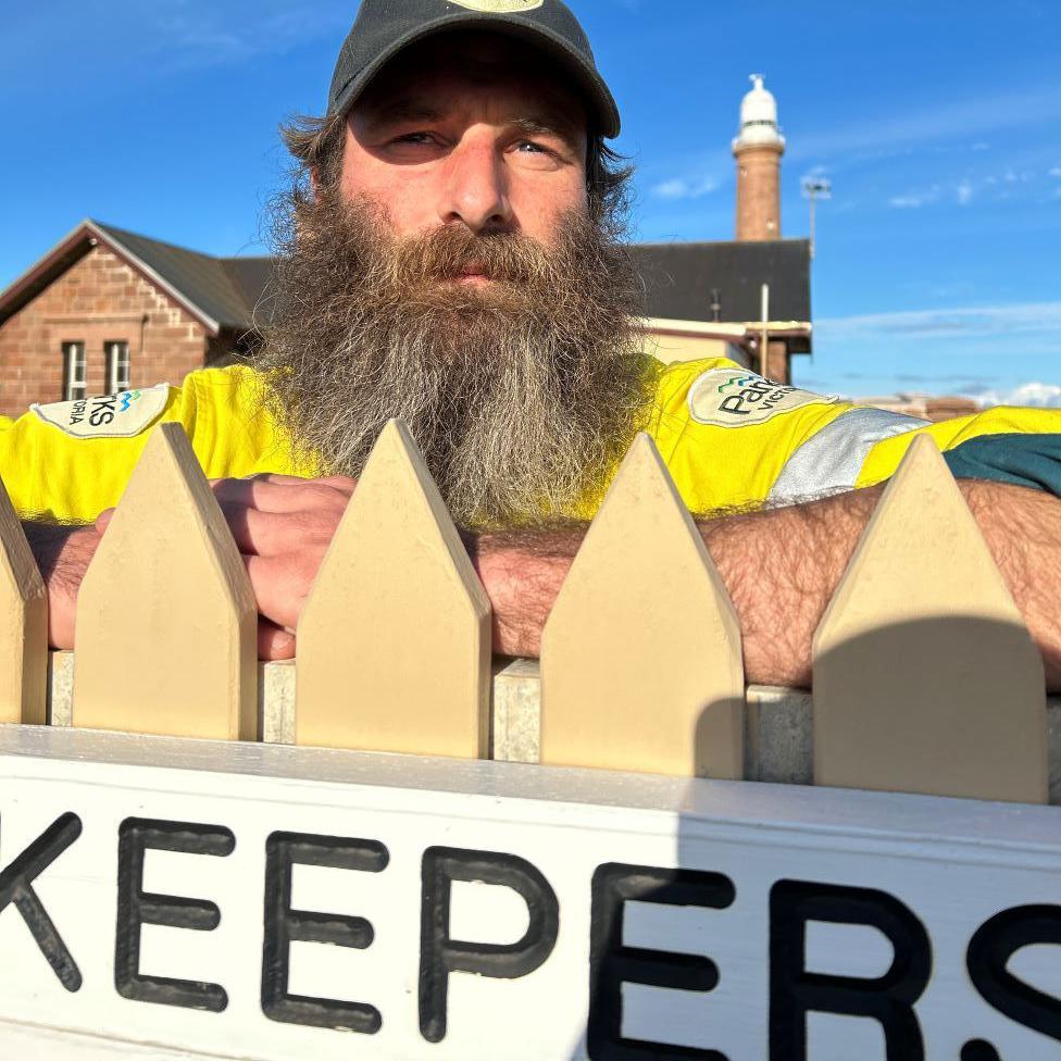 Sandy Duthie, man with large greying beard, yellow Parks Victoria jacket, leaning on a wooden fence with wording which says 'Keepers' with a lighthouse in the background, and blue skies.