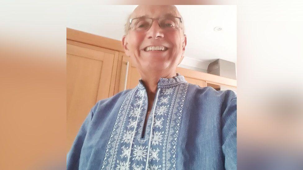 John Eborn, who has grey hair and glasses, smiles as he looks down at the camera. He is wearing traditional Ukrainian dress which was given to him by Michael Velychko. It is a blue, collarless top with a white pattern running down the middle.