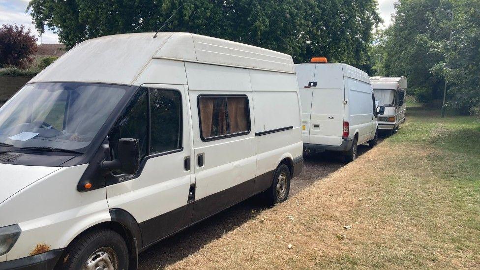 Thre white vans are parked on the side of a road next to grass that has become slightly brown in the sun. There are trees behind the parked vans.