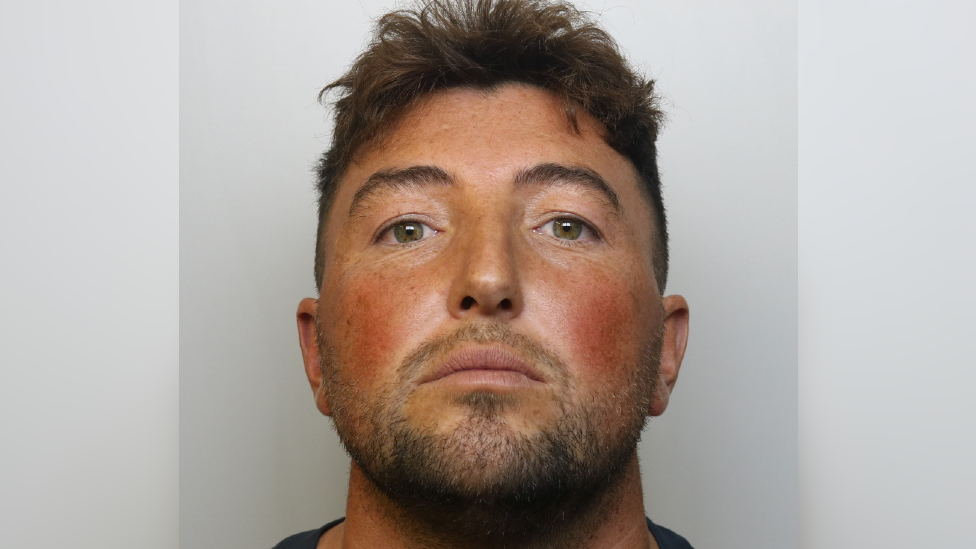 Mugshot of Dominic Capaldi. He has dark, short hair and hazel eyes. He also has a small amount of facial hair. He is pictured against a light grey background, looking directly at the camera with a blank expression. 