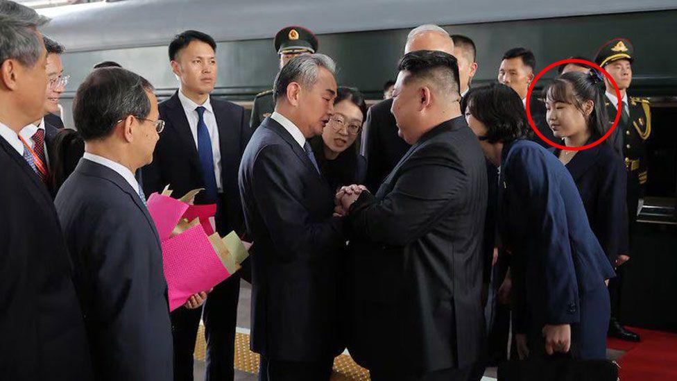 Kim shakes hands an official on the train platform, surrounded by other officials and uniformed guards. His daughter stands behind him, wearing a black dress suit.