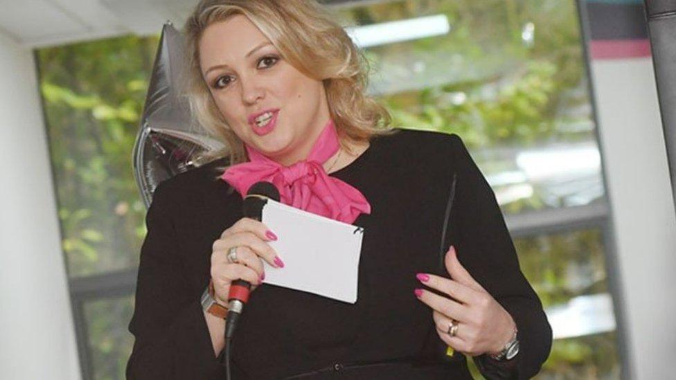 A woman with blonde hair, slightly curly, to her shoulders, holds a microphone in her right hand with a piece of paper in it too. She has a black top on.
