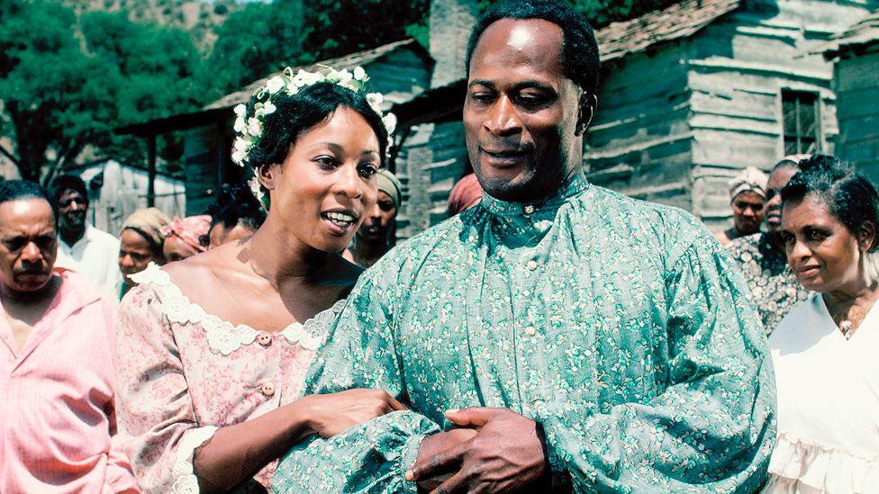 Madge Sinclair and John Amos arm in arm in colourful clothes in a scene from Roots in 1977