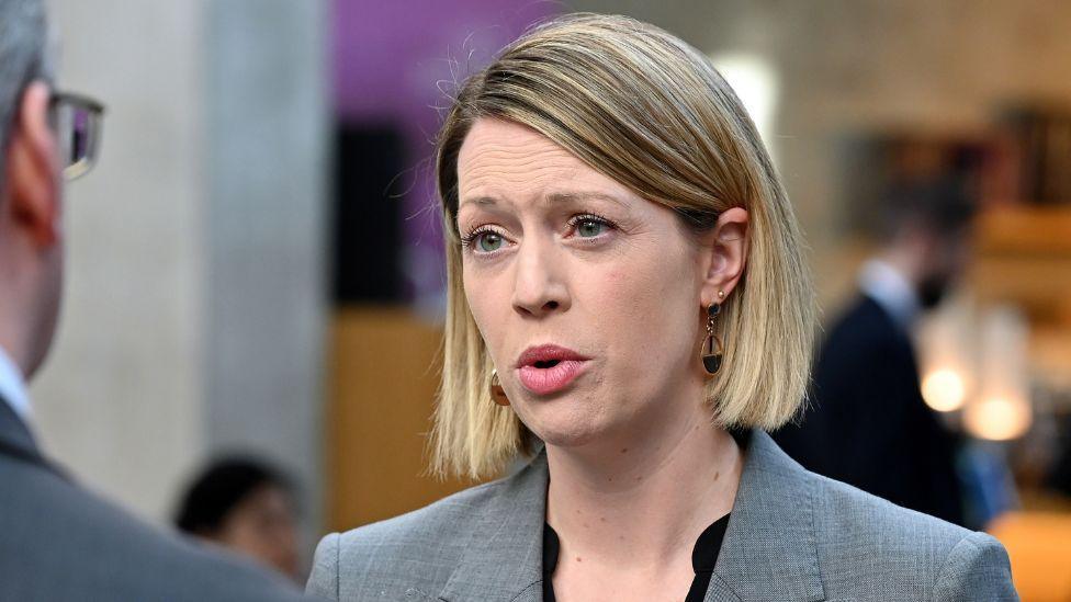 Jenny Gilruth, who has short blonder hair, speaks to a journalist, who has his back to the camera. 
