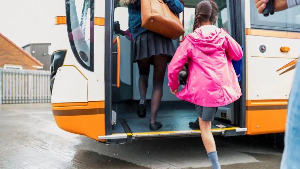 An orange and white school bus with a girl wearing a pink raincoat and grey socks mounting the step at the entrance door. There is a taller girl with a blue top and dark blue skirt ahead of her. She is carrying an orange bag. The bus is parked on grey slabs.