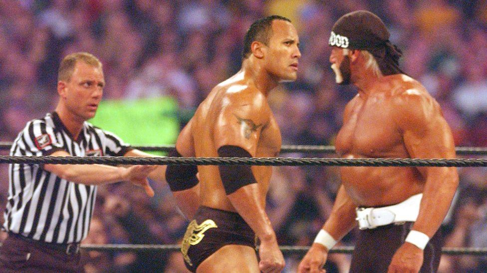 Dwayne Johnson and Hulk Hogan square up, eyeing each other menacingly, dwarfing the referee who's trying to keep order next to them, in the ring at Wrestlemaia X8 in 2002