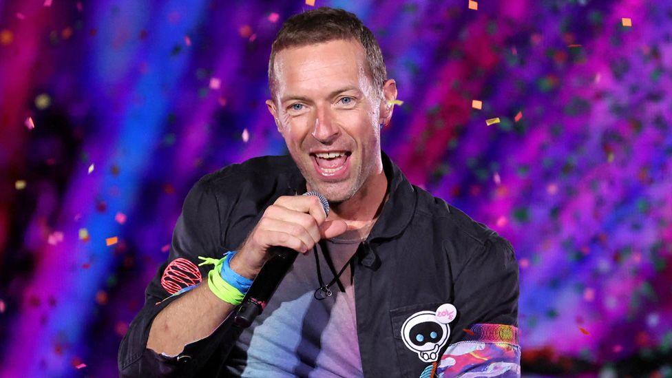 Coldplay frontman Chris Martin singing on stage in front of a blue-purple background and with pieces of confetti falling