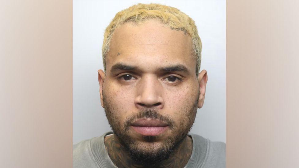 Mug shot of Chris Brown's face showing him with blond hair, short dark brown facial hair and a grey T-shirt
