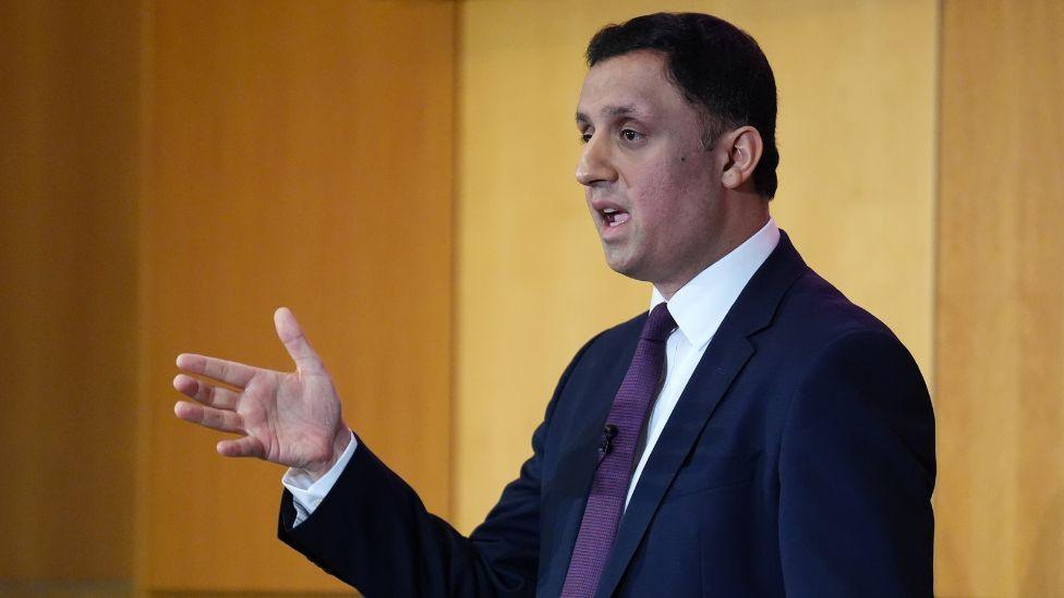 Anas Sarwar, with dark hair and wearing a dark suit, white shirt and purple tie, photographed in a medium close-up. He is standing side on to the camera, with his right hand raised in front of him. 