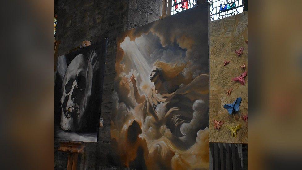 Three paintings depicting different artworks are displayed inside St Cuthbert's Church. There are stained windows in the background.