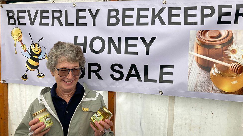 A woman wearing glasses and a green jacket holds up two jars of honey in front of a sign that read Beverley Beekeeper Honey For Sale