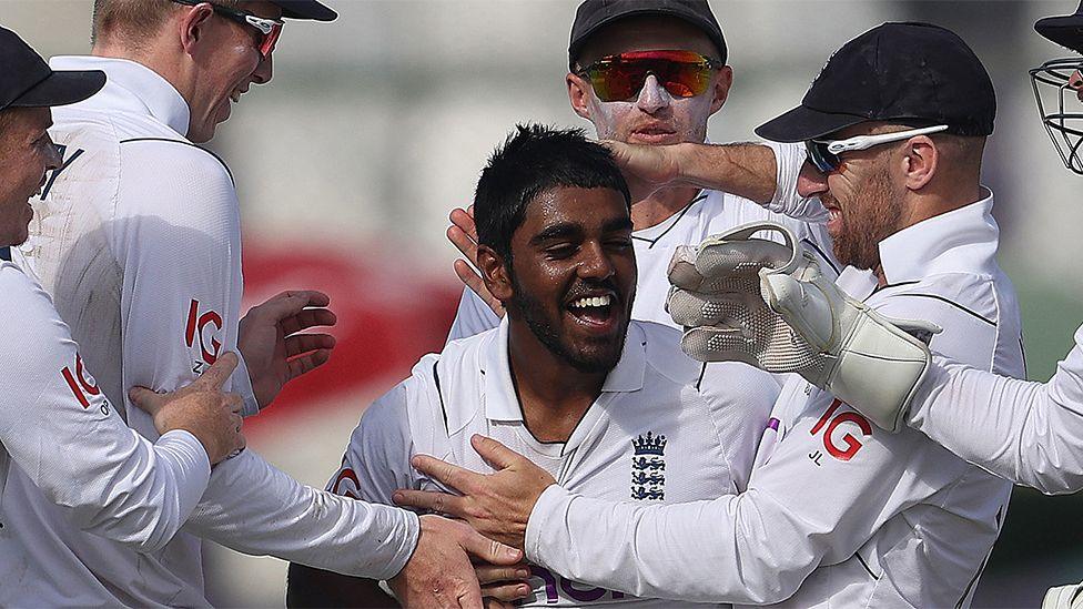 Rehan Ahmed celebrating after taking a wicket against Pakistan in 2022. There are other 5 other players around him congratulating him, patting him on the head and body. They are all wearing the white England jersey