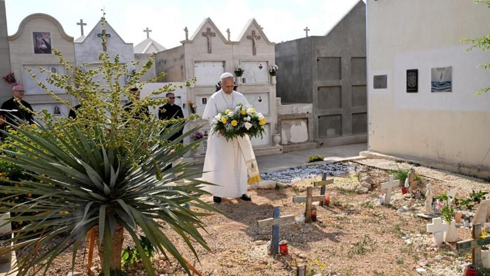 Papa qəbiristanlıqda güllər tutur. Onun arxasında qara kostyumlu rəsmilər və üzərində xaçlar olan ağ binalar sırası var.