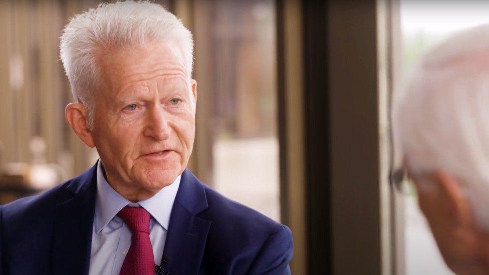 An older man with sharp white hair, wearing a navy blue suit, light blue shirt and crimson tie, is being interviewed by another man with white hair, whose head we see the back of.