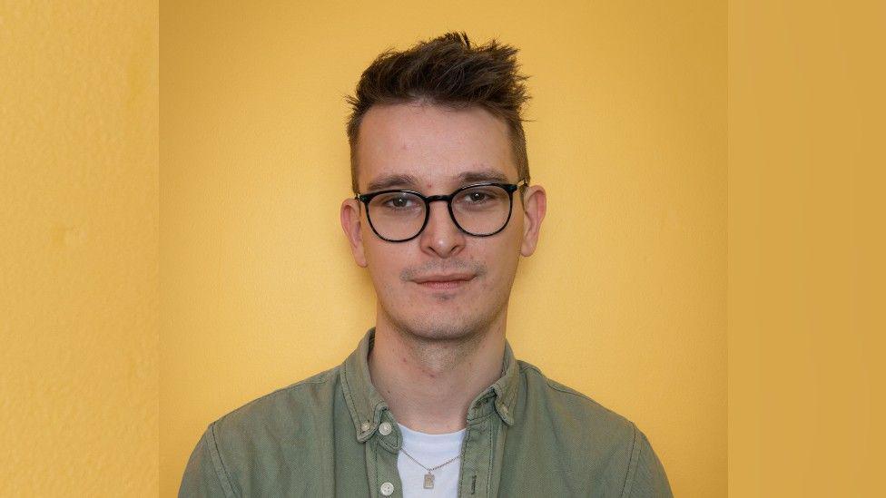 Matt Justice standing against a yellow wall. He is wearing a green shirt, white t-shirt and black framed round glasses. He has short brown hair which sticks up and is smiling faintly at the camera. 